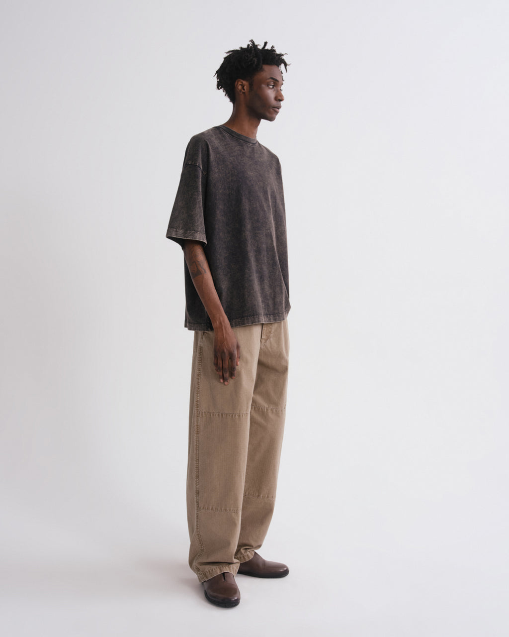 A model stands in profile wearing a washed charcoal gray oversized t-shirt, beige wide-leg cargo pants, and brown leather shoes against a plain white studio background.