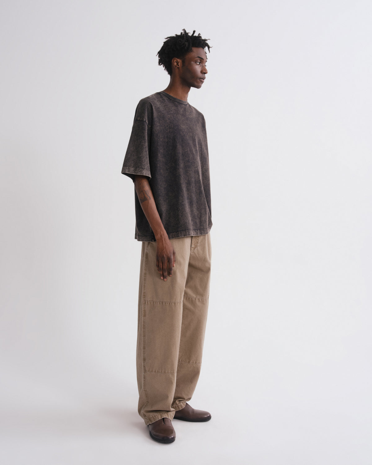 A model stands in profile wearing a washed charcoal gray oversized t-shirt, beige wide-leg cargo pants, and brown leather shoes against a plain white studio background.