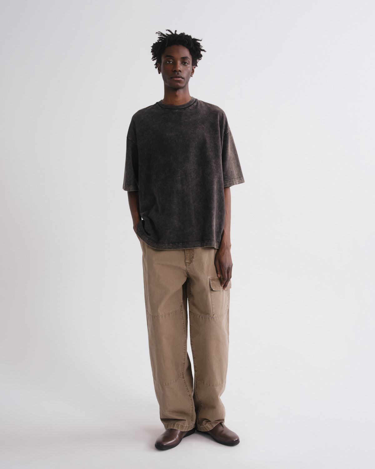A man stands against a plain light gray studio background wearing an oversized dark charcoal washed t-shirt, beige cargo pants, and brown shoes.