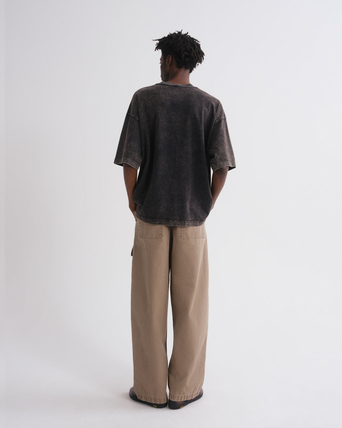A man is shown from behind wearing a dark gray mineral-washed t-shirt and beige wide-leg trousers, standing against a plain white studio background.