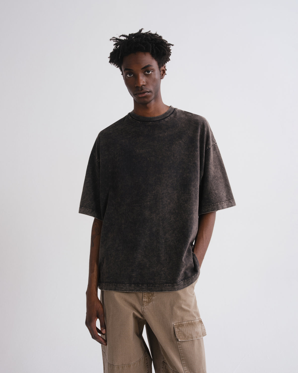 A man wearing a dark brown acid-wash short-sleeve t-shirt and beige cargo pants, standing against a light gray studio background.
