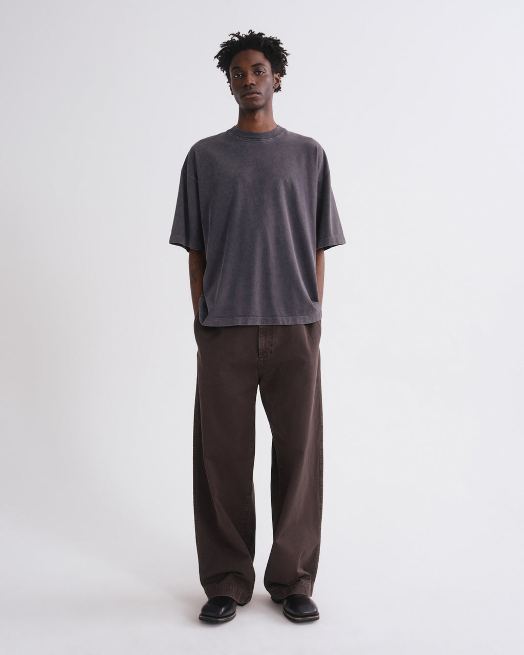 A man stands against a plain light gray studio background wearing an oversized dark charcoal gray t-shirt and loose dark brown pants.