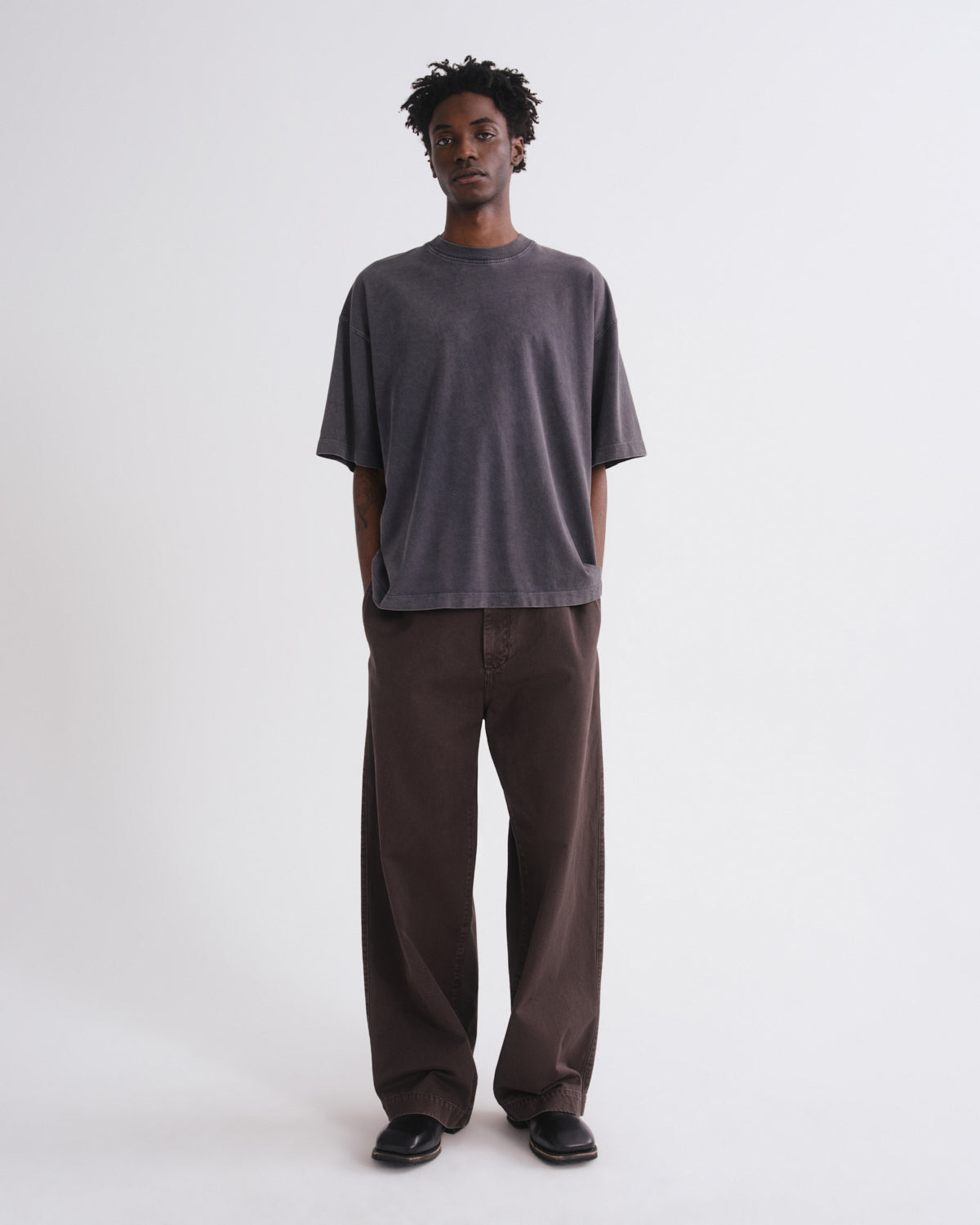 A man stands against a plain light gray studio background wearing an oversized dark charcoal gray t-shirt and loose dark brown pants.