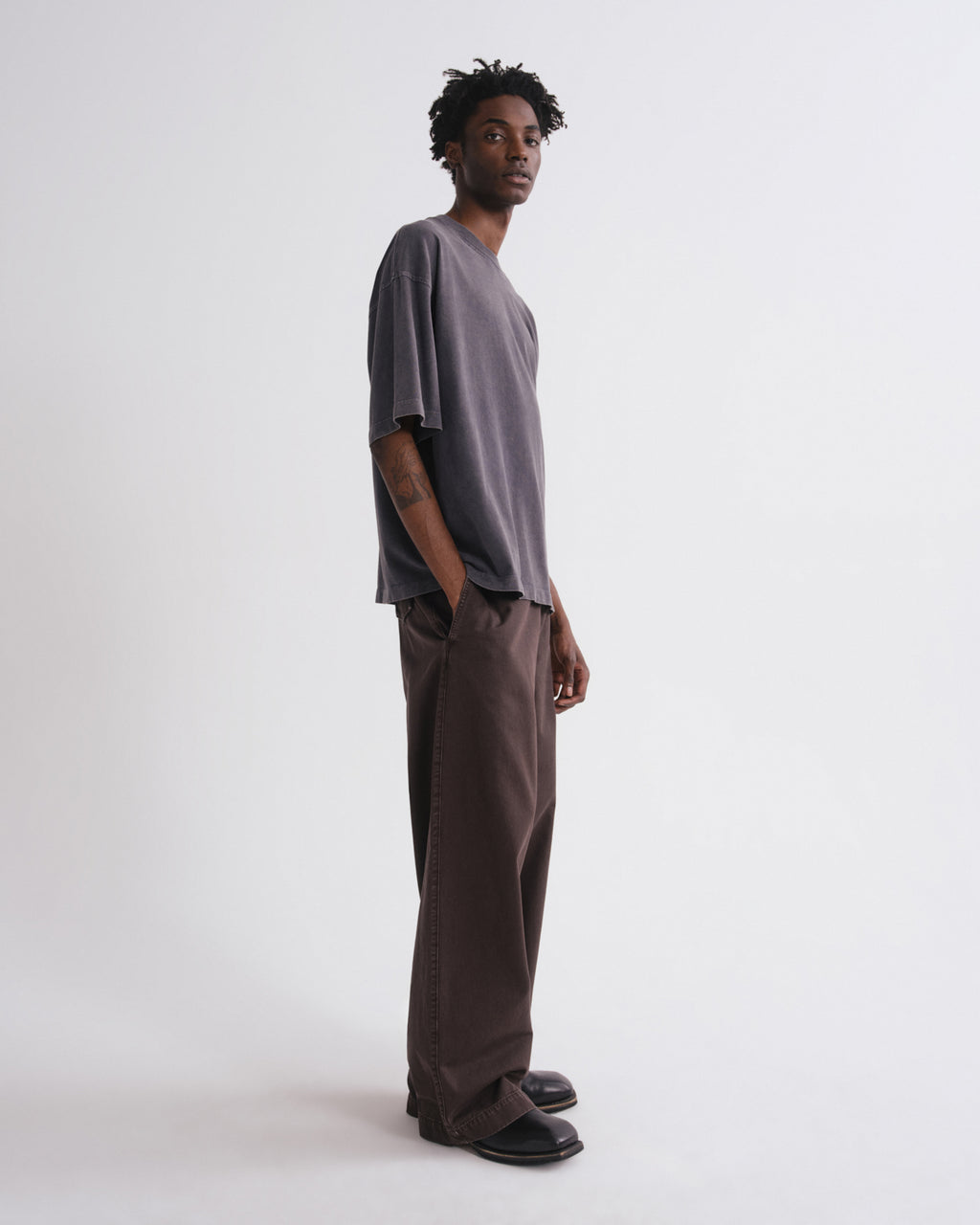 A man wearing a loose, oversized short-sleeve t-shirt in a muted grayish-purple and matching wide-leg trousers, standing in profile against a plain light gray studio background.