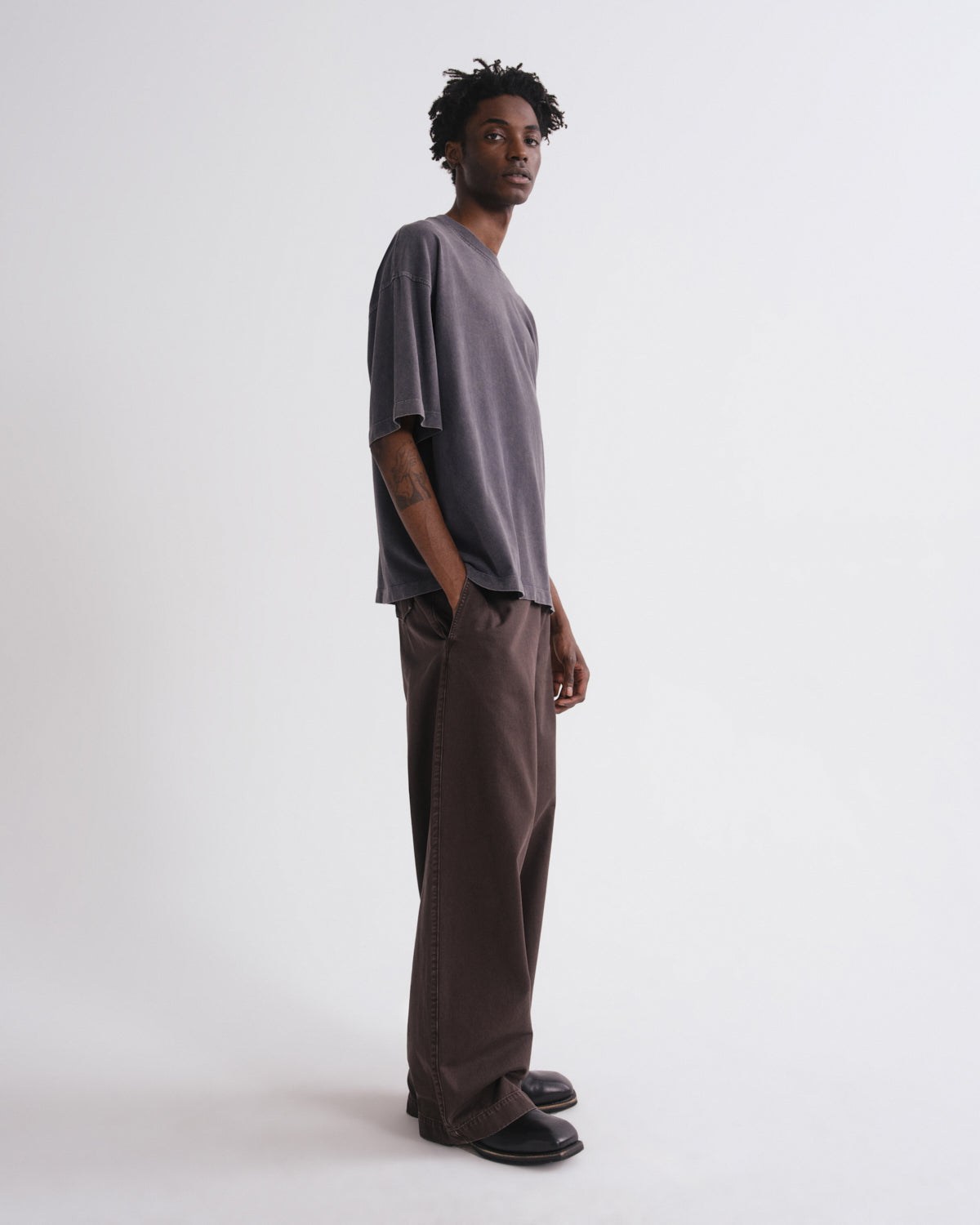A man wearing a loose, oversized short-sleeve t-shirt in a muted grayish-purple and matching wide-leg trousers, standing in profile against a plain light gray studio background.