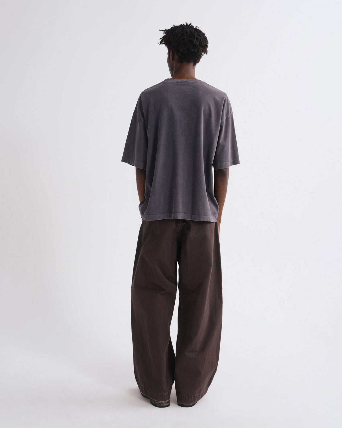 A person shown from the back wearing an oversized dark gray short-sleeve t-shirt and very wide-leg dark brown pants, standing against a plain light gray studio background.