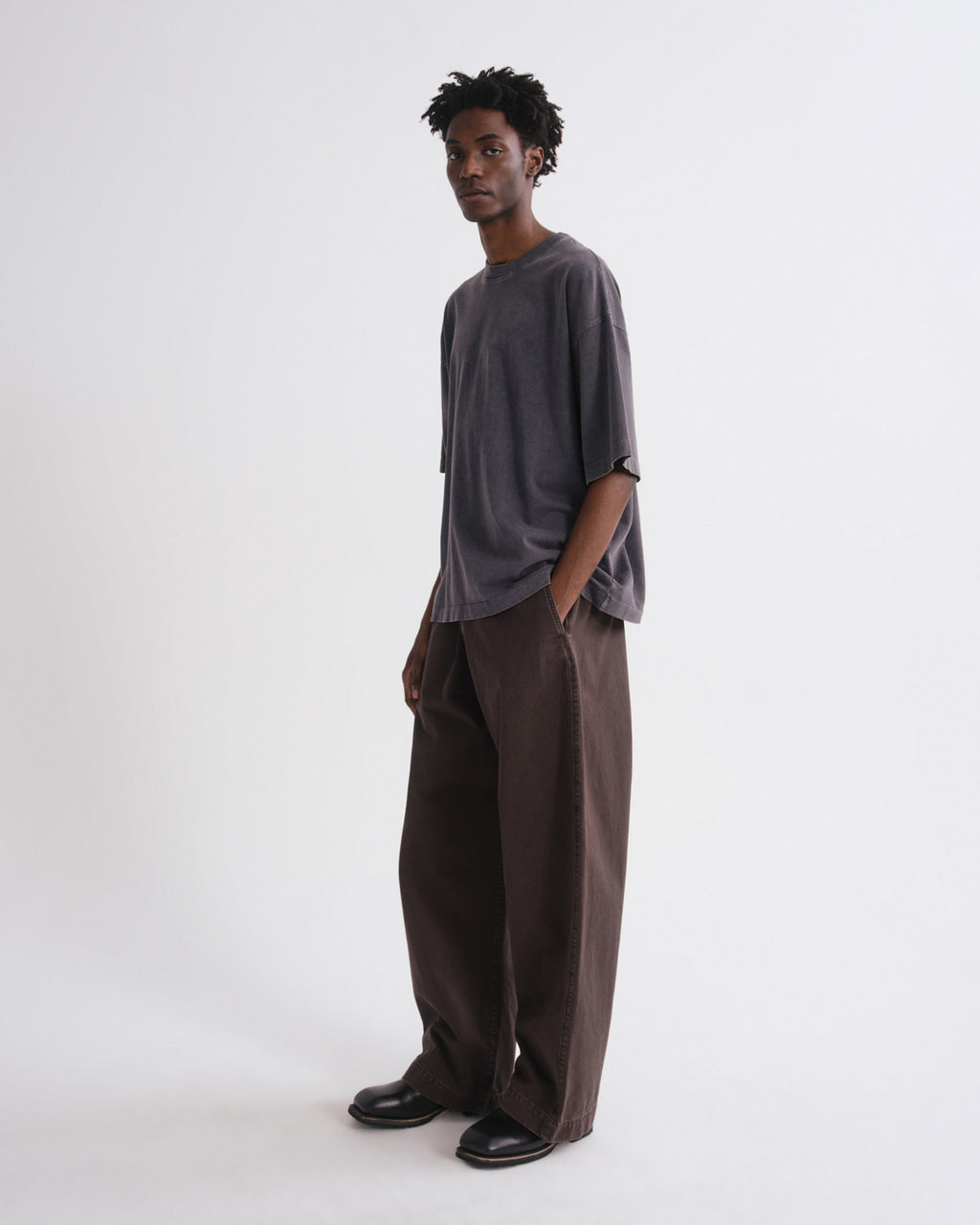 A man modeling a loose-fit dark gray oversized t-shirt and matching dark brown wide-leg trousers, standing against a plain light gray studio background.