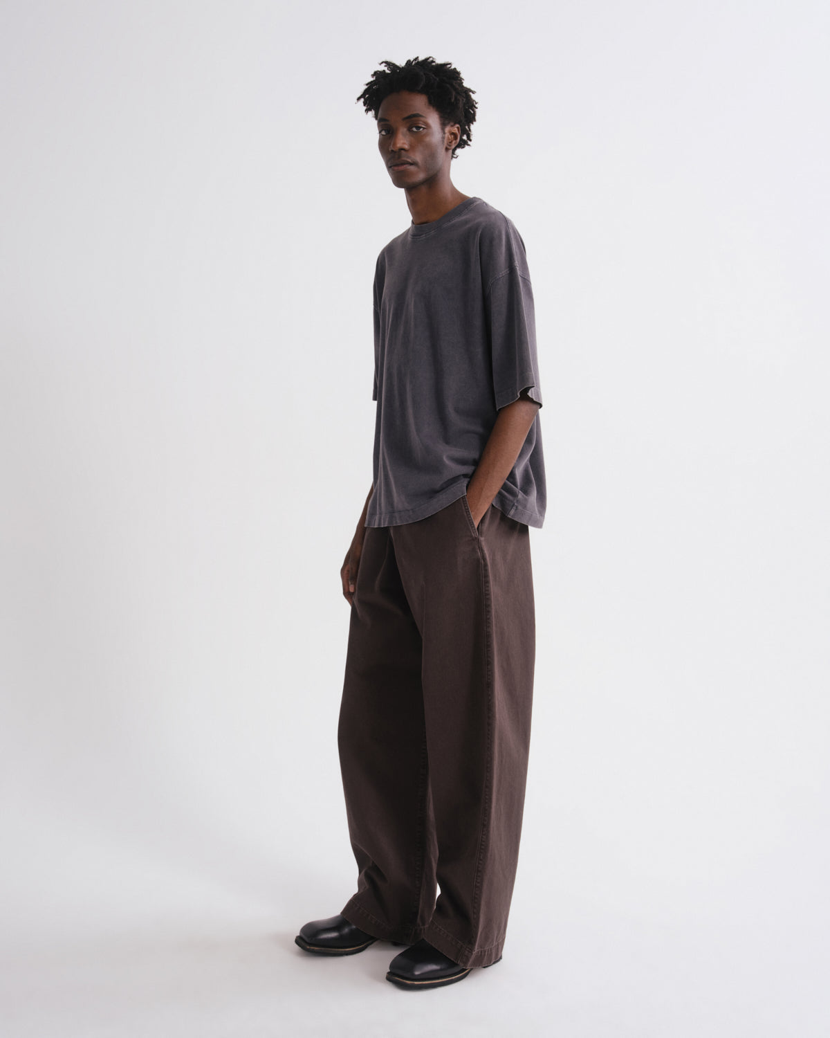 A man modeling a loose-fit dark gray oversized t-shirt and matching dark brown wide-leg trousers, standing against a plain light gray studio background.