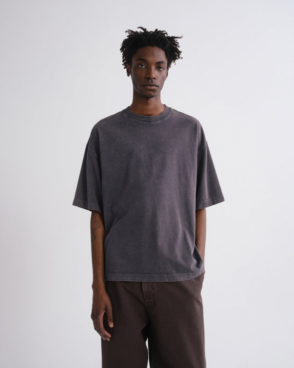 A man wearing an oversized dark gray washed t-shirt and brown pants, standing against a plain light gray studio background.