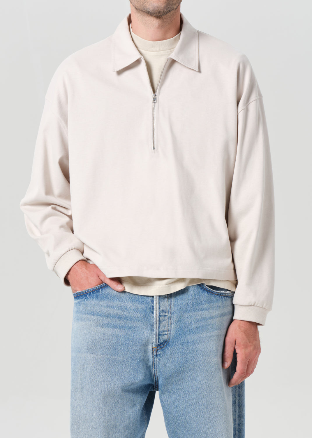 Person wearing a beige zip-up sweatshirt and light blue jeans on a plain background