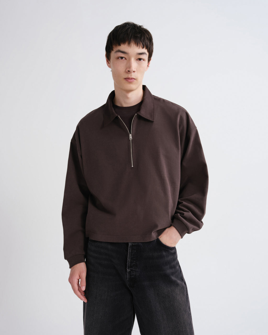 Close up of a person wearing a brown shirt and black pants standing against a grey background