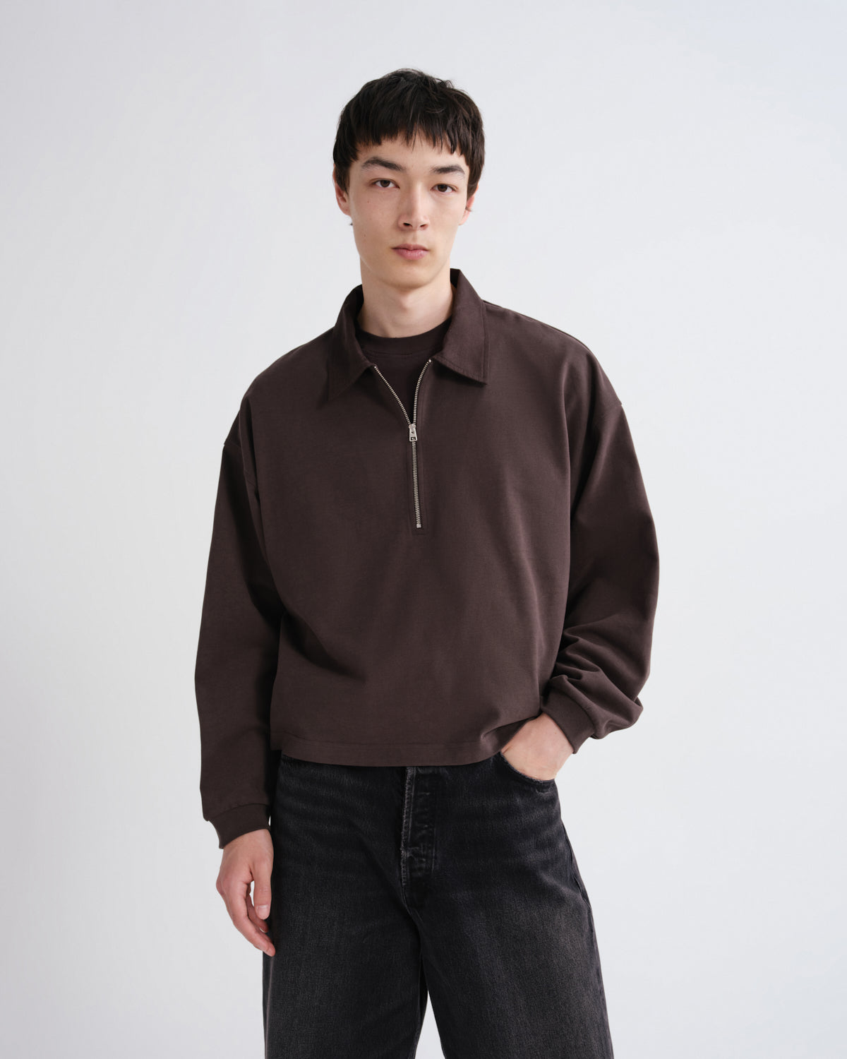 Close up of a person wearing a brown shirt and black pants standing against a grey background