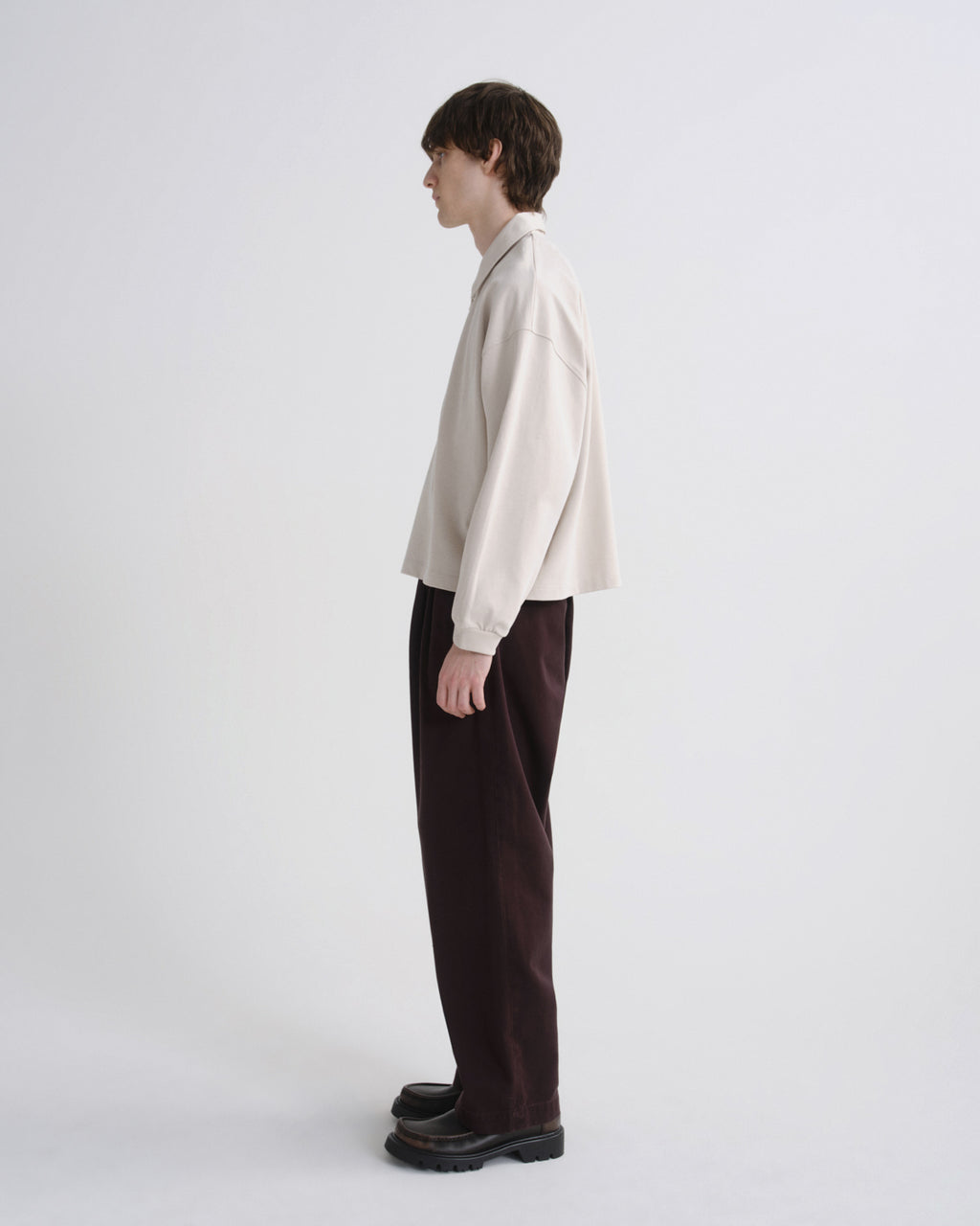 A model shown in profile wearing a cream-colored oversized long-sleeve shirt and dark brown wide-leg trousers, standing against a plain light gray studio background.