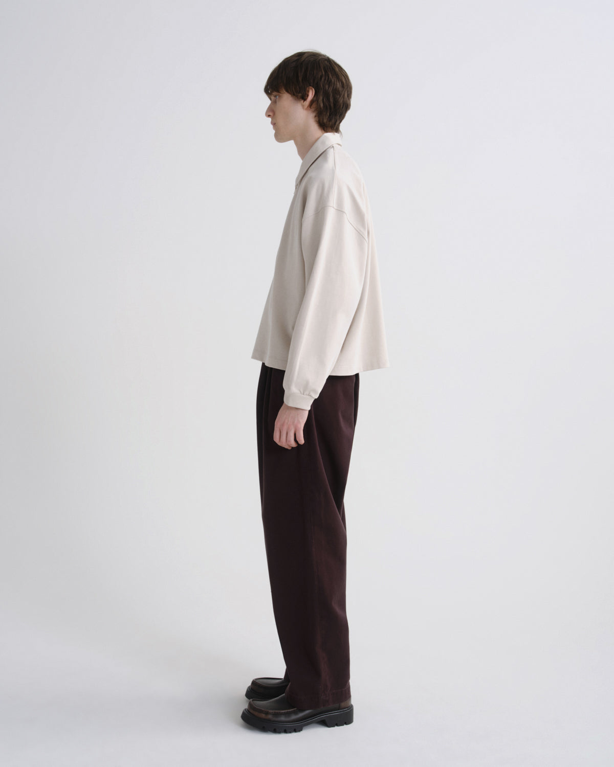 A model shown in profile wearing a cream-colored oversized long-sleeve shirt and dark brown wide-leg trousers, standing against a plain light gray studio background.