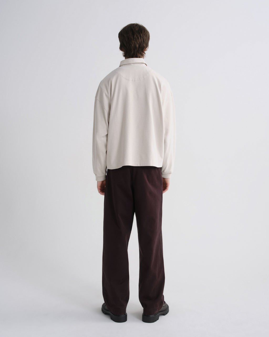 A man viewed from behind wearing a cream-colored long-sleeve collared shirt and dark brown trousers, standing against a plain light gray studio background.