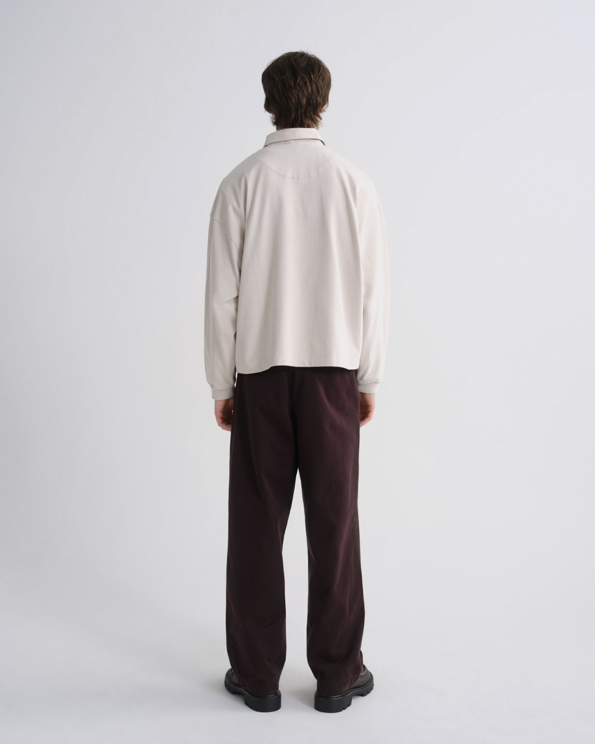 A man viewed from behind wearing a cream-colored long-sleeve collared shirt and dark brown trousers, standing against a plain light gray studio background.