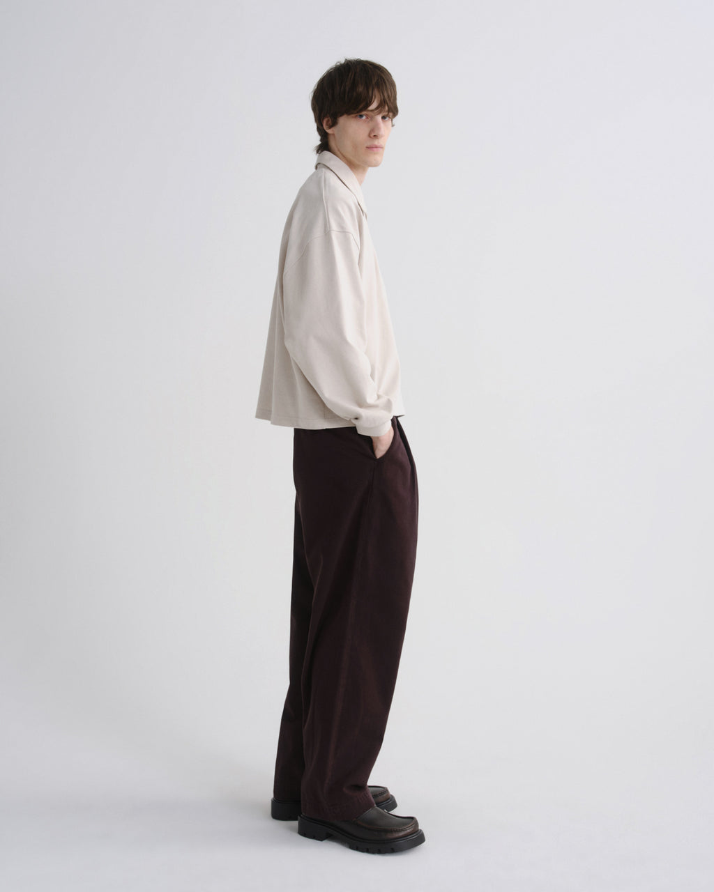 A model stands in profile wearing a cream-colored long-sleeve button-up shirt, dark brown wide-leg trousers, and black loafers against a plain light gray studio background.