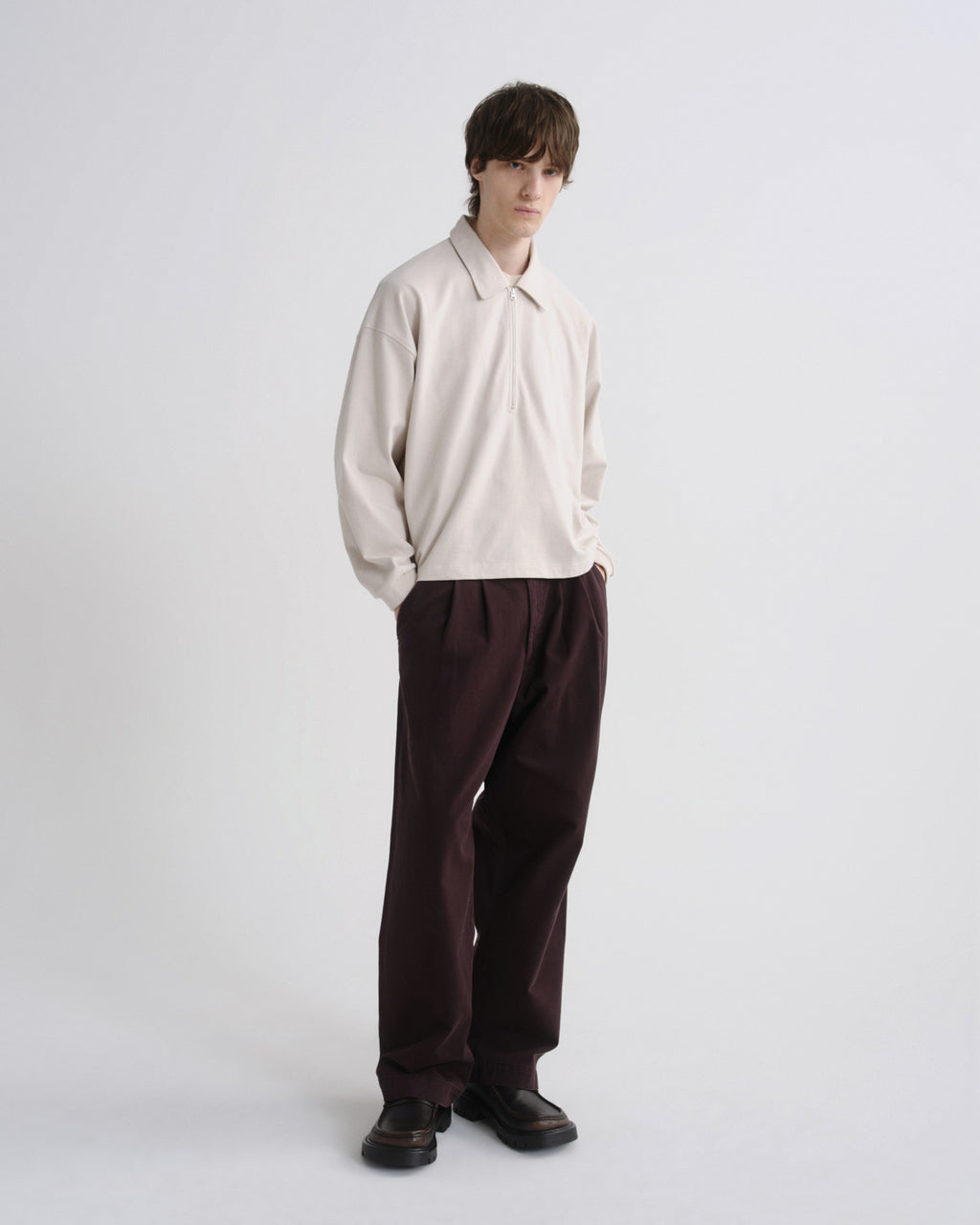 A model stands against a light gray studio background wearing a cream long-sleeve collared shirt and dark brown pleated trousers.