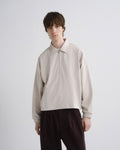 A model wearing a light beige long-sleeve half-zip pullover with a collar, paired with dark brown trousers, against a plain light gray background.