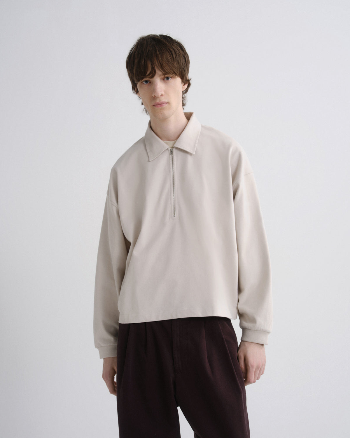 A model wearing a light beige long-sleeve half-zip pullover with a collar, paired with dark brown trousers, against a plain light gray background.