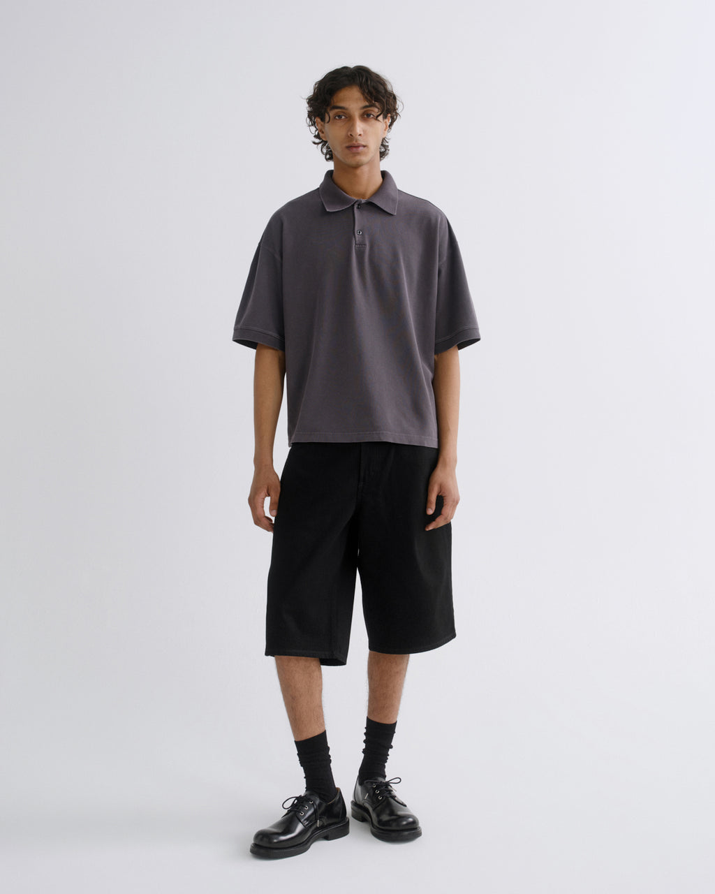 A model stands against a light gray studio background wearing a dark gray short-sleeve polo shirt, black shorts, black socks, and black leather shoes.