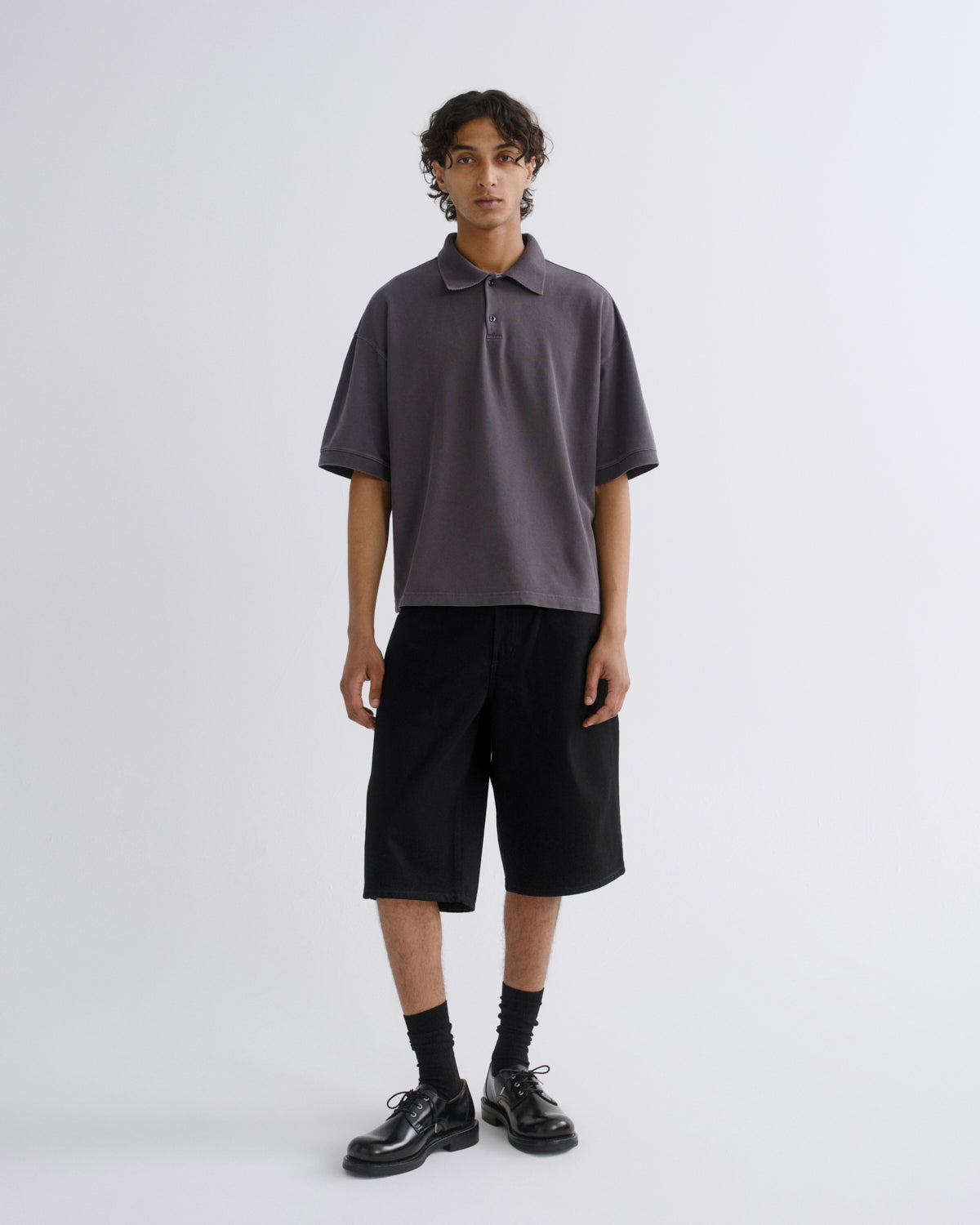 A model stands against a light gray studio background wearing a dark gray short-sleeve polo shirt, black shorts, black socks, and black leather shoes.
