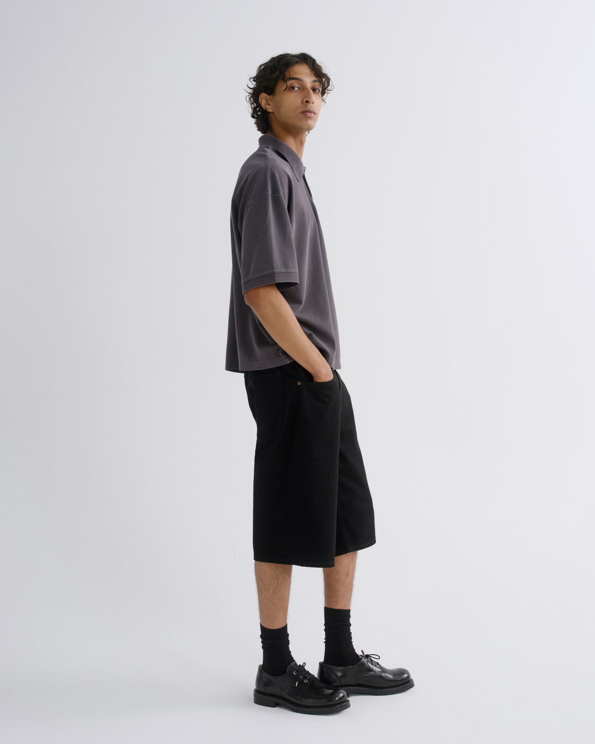 A model stands in profile wearing a dark gray short-sleeve polo shirt, black knee-length shorts, black socks, and black leather shoes against a light gray studio background.