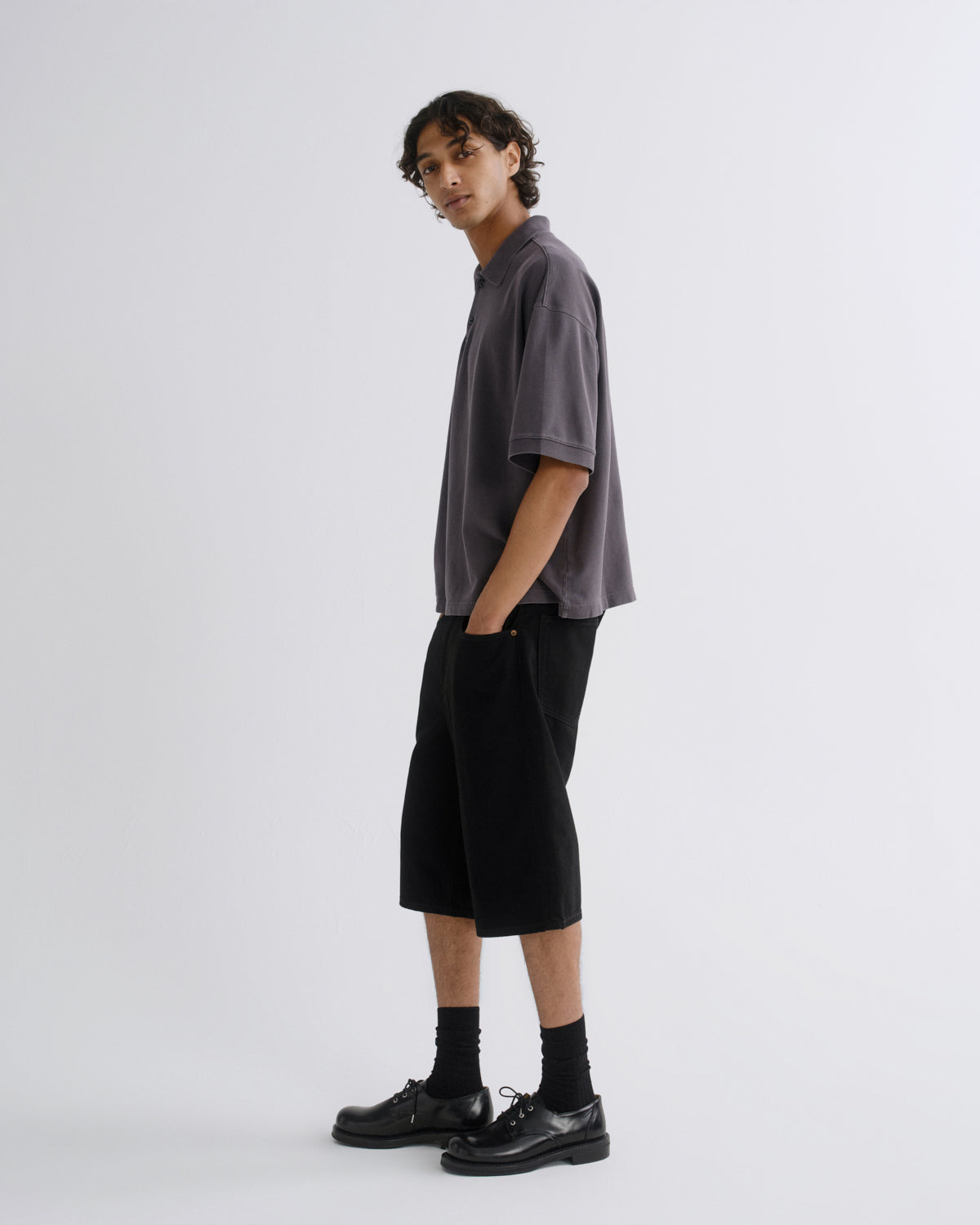 A model stands in profile wearing a dark gray short-sleeve collared shirt, black cropped shorts, black socks, and black leather shoes against a light gray studio background.