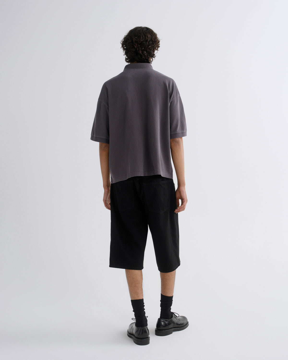 A model shown from the back wearing a dark gray short-sleeve polo shirt, black cropped shorts, black socks, and black leather shoes against a light gray studio background.