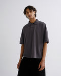 A man wearing a dark gray short-sleeve polo shirt and black pants, standing against a light gray studio background.