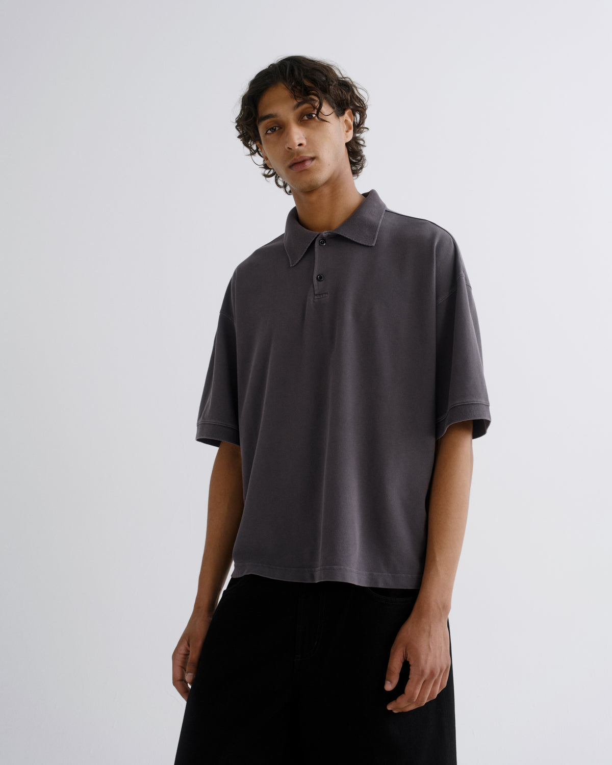 A man wearing a dark gray short-sleeve polo shirt and black pants, standing against a light gray studio background.