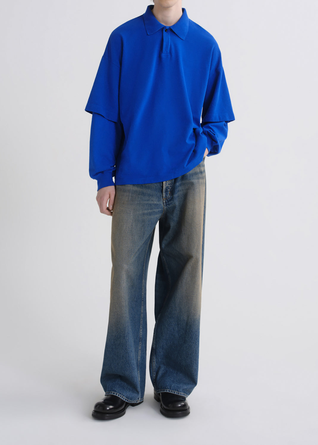 A model wearing a vibrant royal blue long-sleeve collared shirt with layered sleeves and wide-leg faded blue jeans, standing against a grey background.