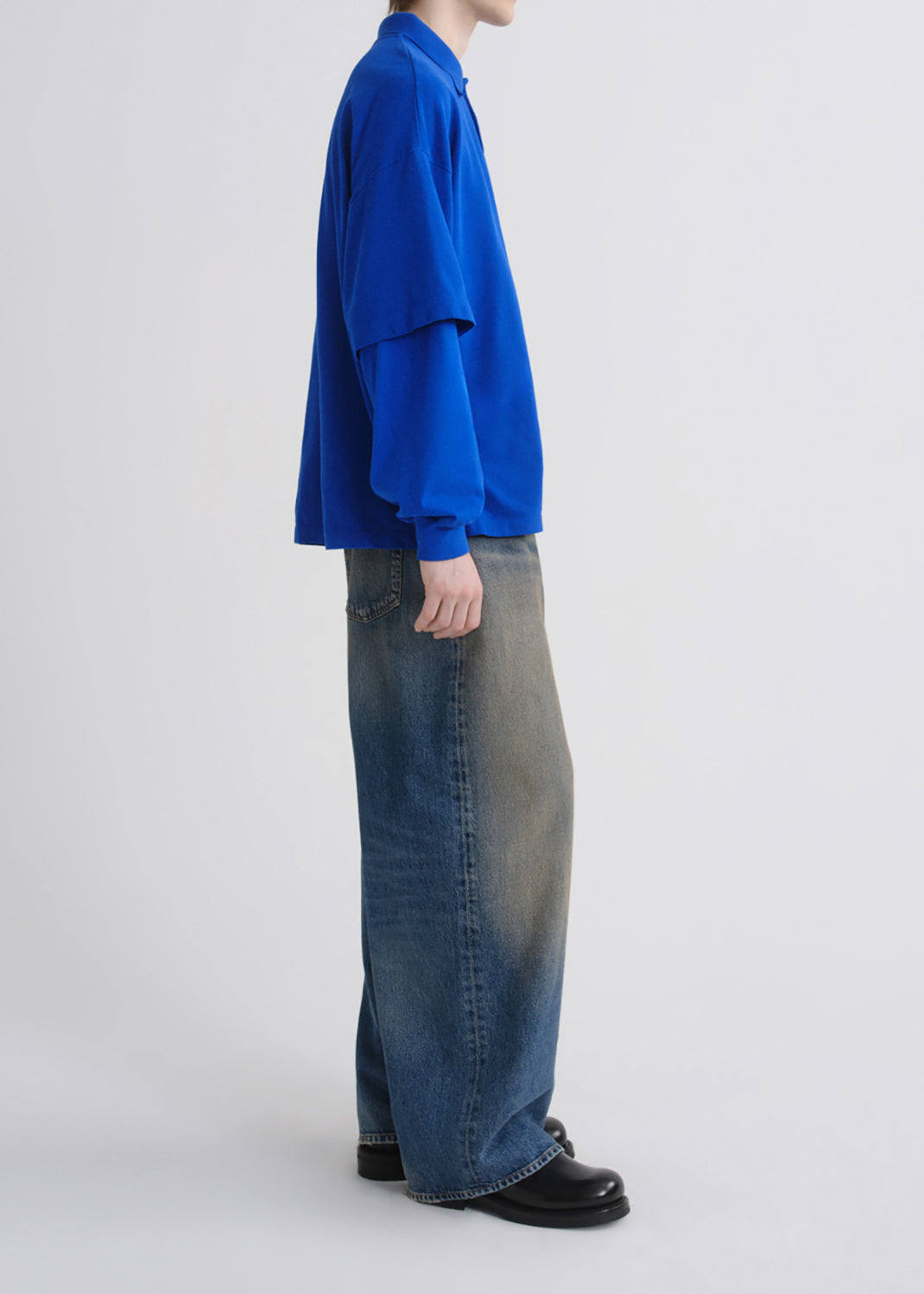 A model shown from the side wearing a vibrant blue long-sleeve collared shirt, wide-leg faded blue jeans, and black leather shoes against a grey background.