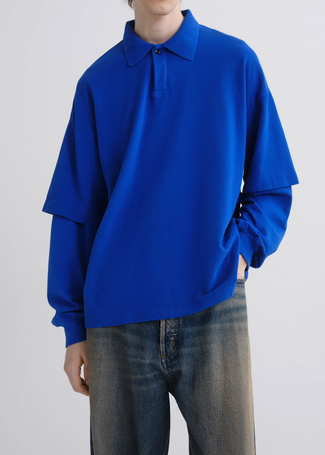 A person wearing a bright royal blue long-sleeve polo shirt with a relaxed oversized fit and dark blue jeans, photographed against a grey background.
