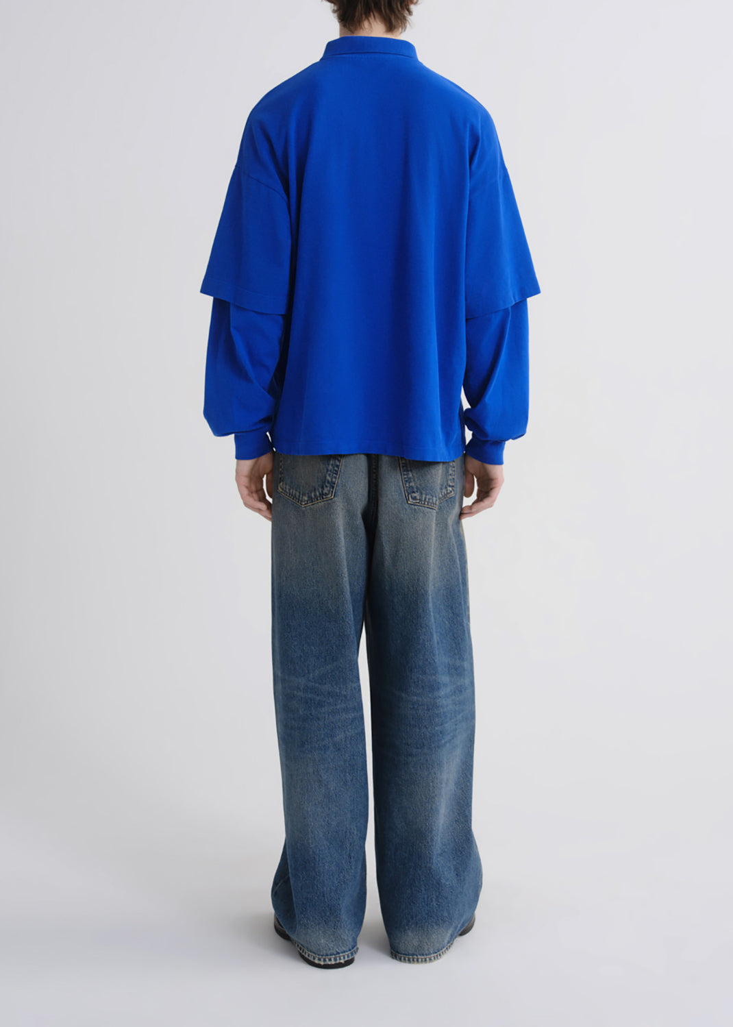 A model viewed from the back wearing a bright royal blue long-sleeve layered shirt and wide-leg blue jeans against a grey background.