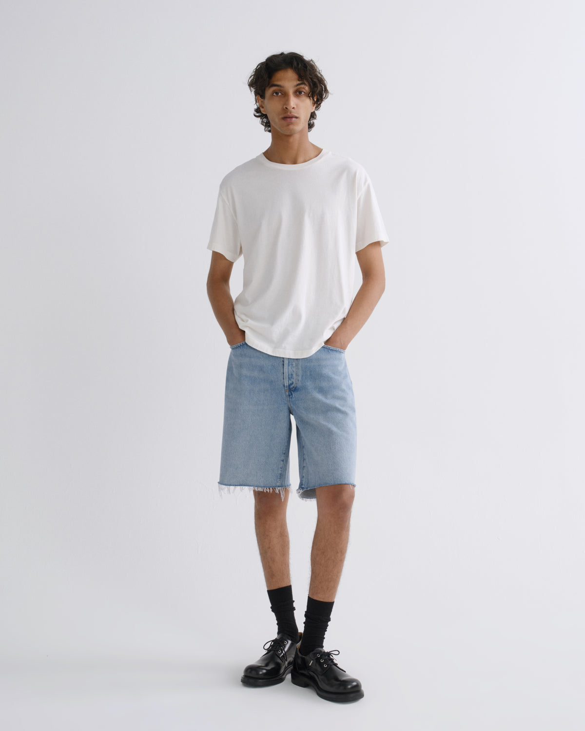 A model stands against a light gray studio background wearing a white crew-neck t-shirt, light-wash denim shorts with raw hems, black socks, and black leather shoes.