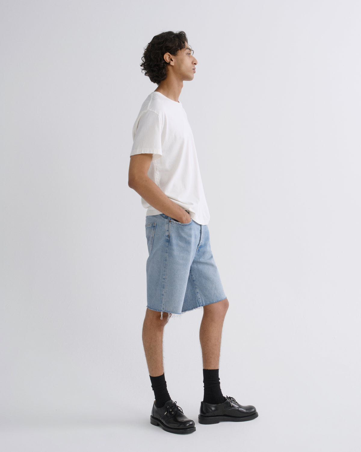 A man stands in profile wearing a white short-sleeve T-shirt, light-wash denim shorts, black socks, and black leather shoes against a clean light gray studio background.