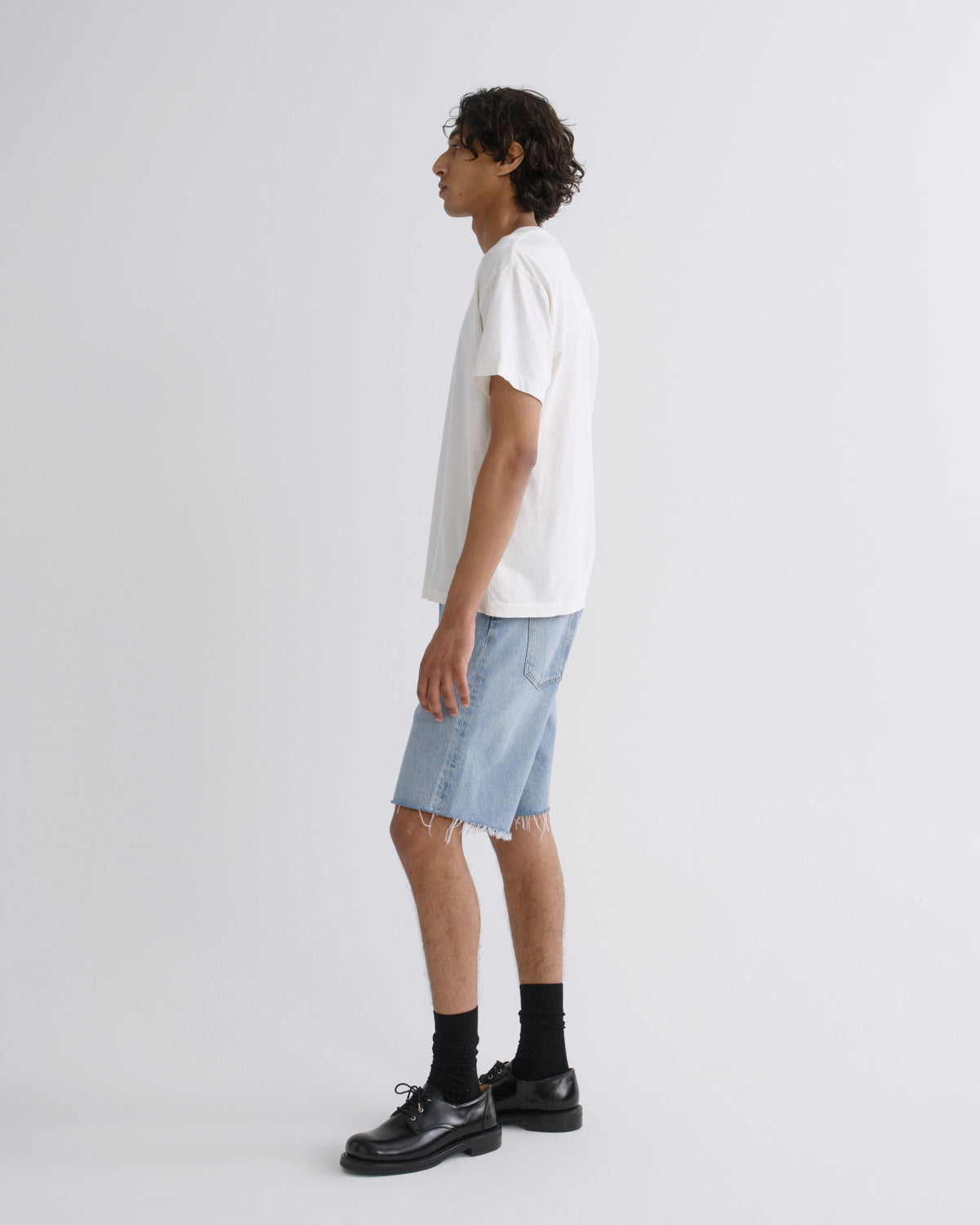 A man shown in profile wearing a white short-sleeve t-shirt, light-wash denim shorts, black socks, and black leather shoes against a plain light gray studio background.