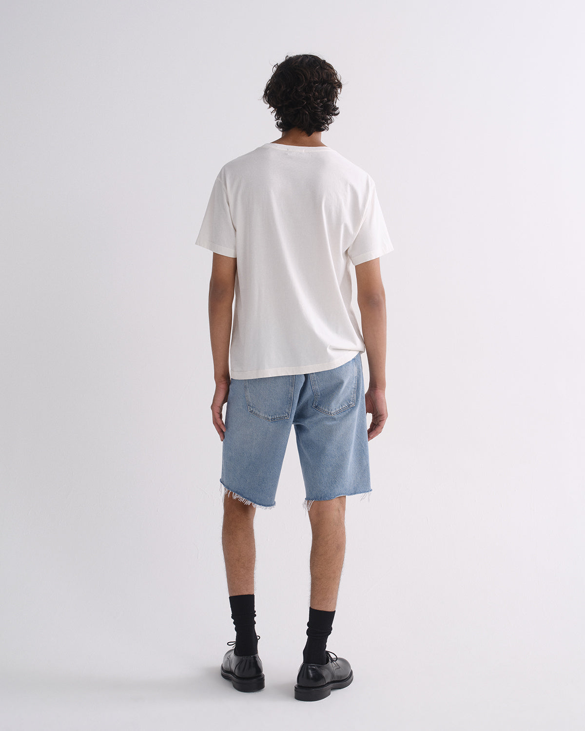 A man viewed from behind wearing a white crewneck t-shirt, light blue denim shorts with raw hems, black socks, and black leather shoes, standing against a light gray studio background.