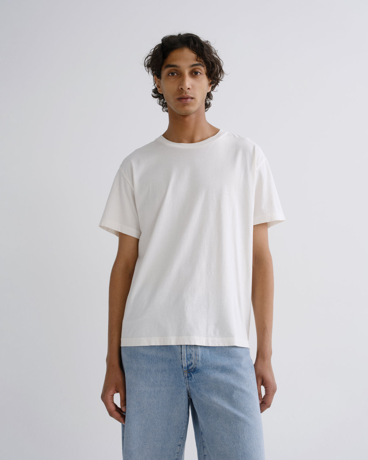 A man wearing a plain white short-sleeve crewneck t-shirt and light blue jeans, standing against a light gray studio background.