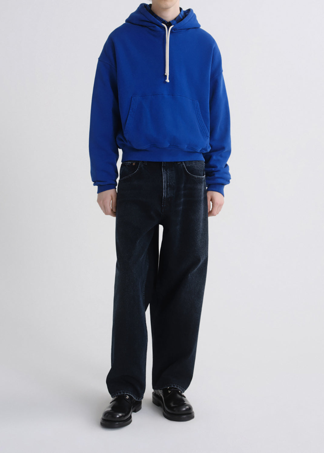 A model wearing a vibrant royal blue pullover hoodie with a white drawstring and dark wide-leg jeans, standing against a grey background.