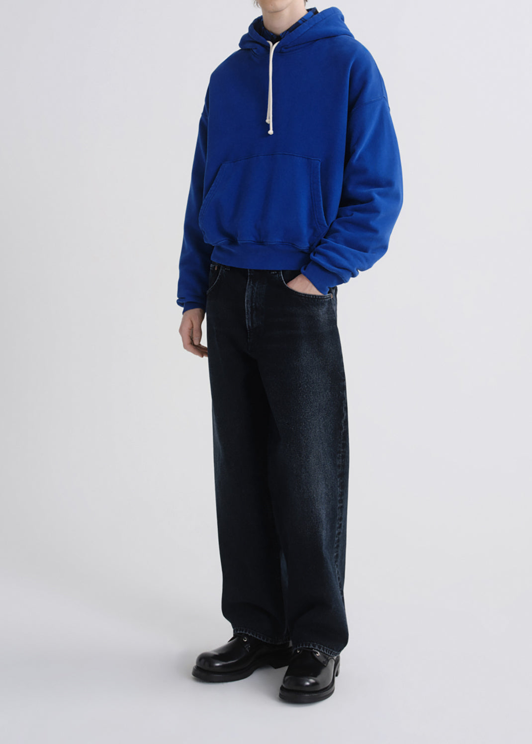 A model wearing a vibrant royal blue oversized pullover hoodie with white drawstrings, paired with dark wide-leg jeans and black leather shoes, standing against a grey background.