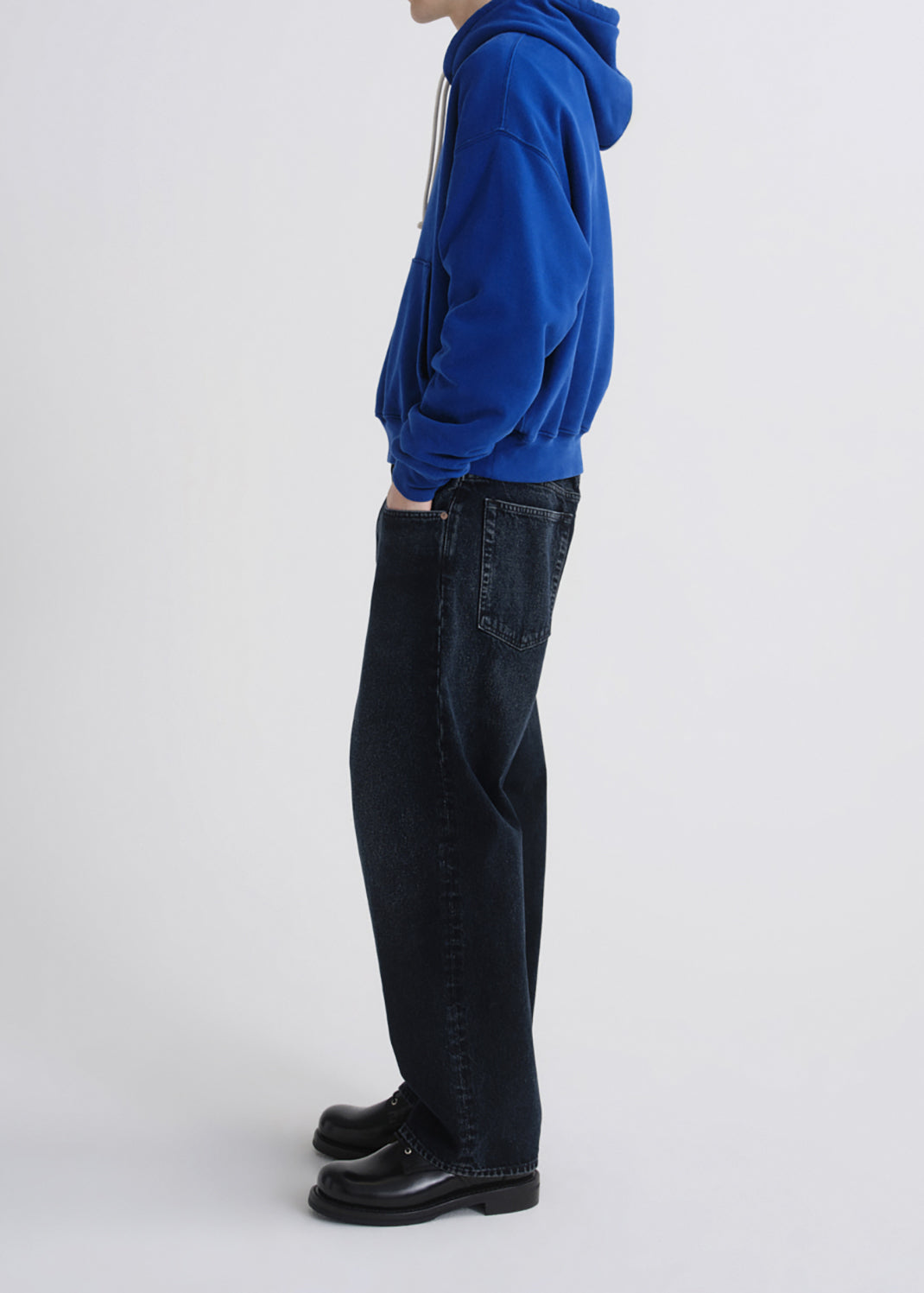 A side-profile view of a person wearing a bright royal blue hoodie, dark indigo jeans, and black leather shoes against a grey background.