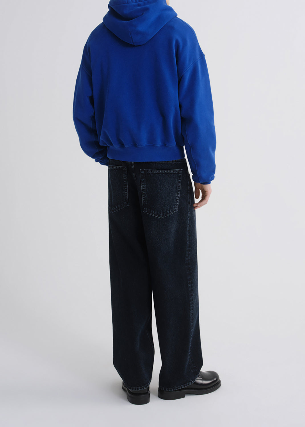 A person shown from the back wearing a bright royal blue cropped hoodie and dark wide-leg jeans, standing against a grey background.