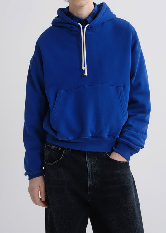 A person wearing a bright royal blue pullover hoodie with white drawstrings and black jeans, photographed against a grey background.