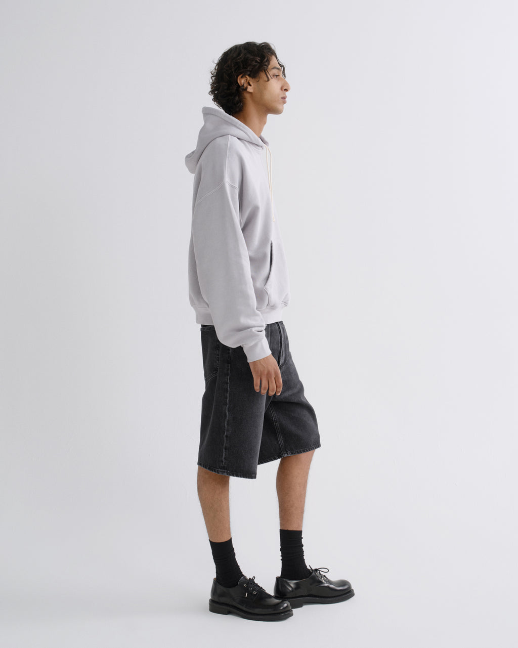 A model shown in profile wearing a light gray pullover hoodie, dark gray denim shorts, black socks, and black leather shoes against a plain light gray studio background.