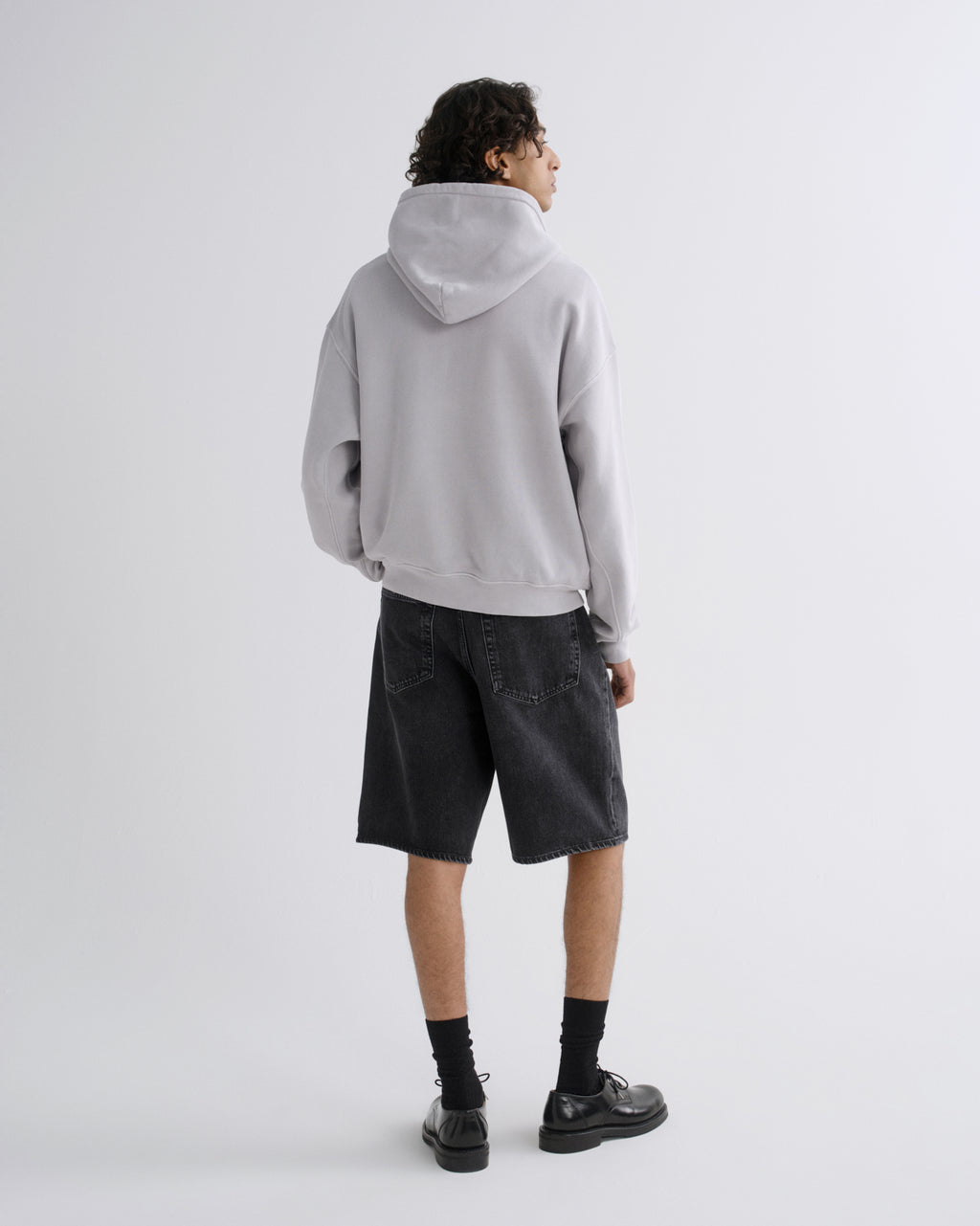 A model shown from the back wearing a light gray oversized hoodie, dark gray denim shorts, black socks, and black leather shoes, standing against a plain light gray studio background.