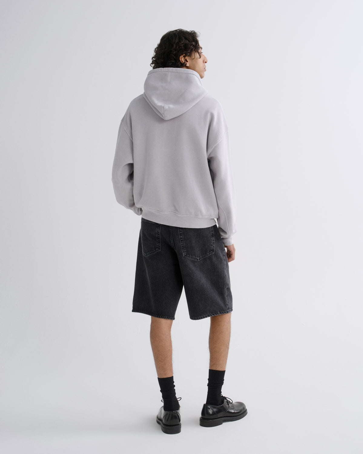 A model shown from the back wearing a light gray oversized hoodie, dark gray denim shorts, black socks, and black leather shoes, standing against a plain light gray studio background.