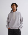 A man wearing a light gray oversized pullover hoodie with cream-colored drawstrings and a front kangaroo pocket, paired with dark jeans, standing against a plain light gray studio background.