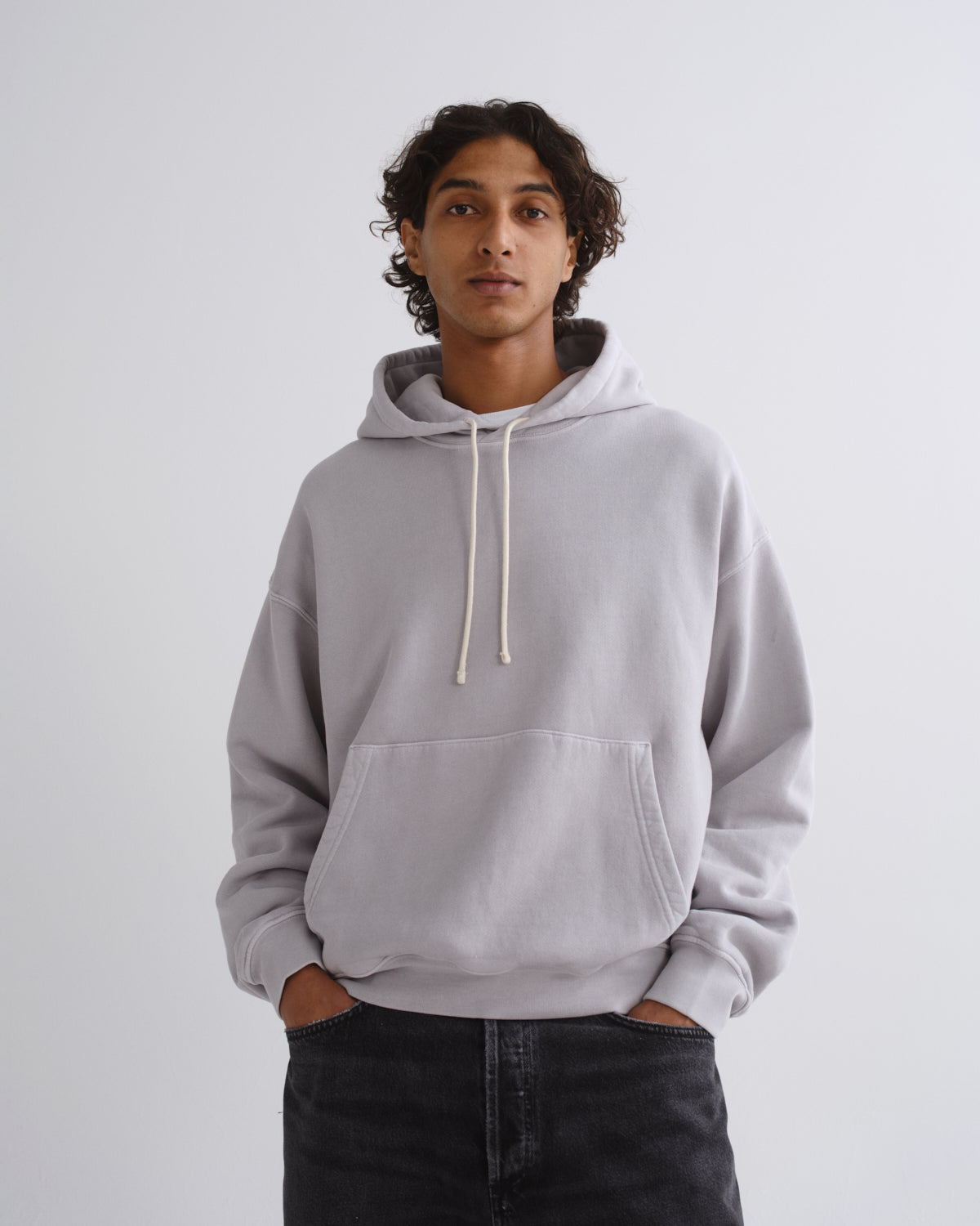A man wearing a light gray oversized pullover hoodie with cream-colored drawstrings and a front kangaroo pocket, paired with dark jeans, standing against a plain light gray studio background.